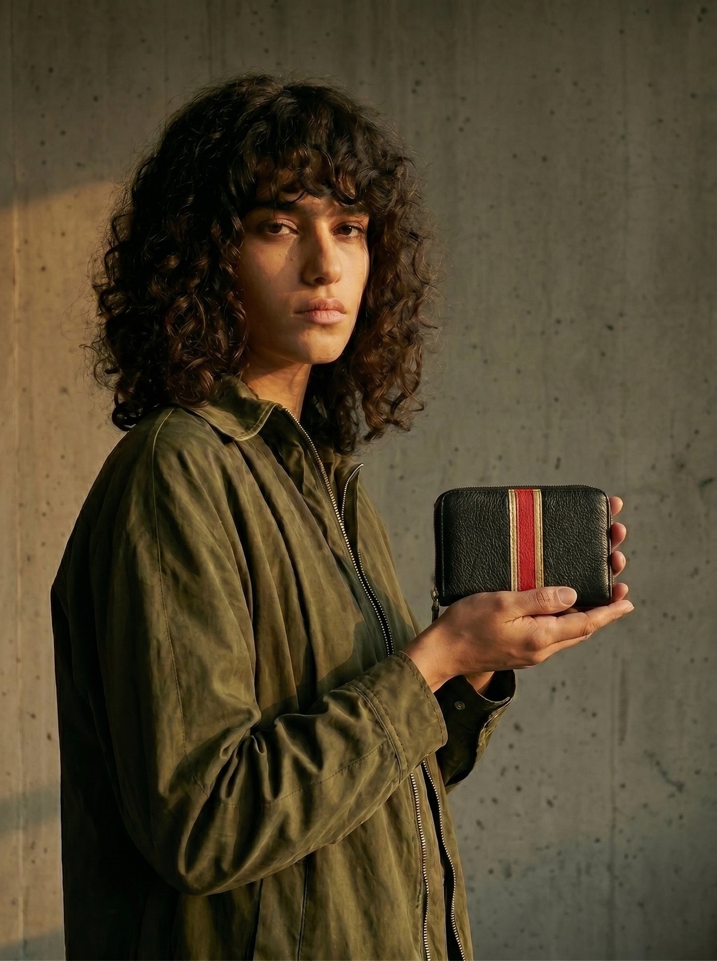 Une jeune femme brune aux cheveux bouclés montre un portefeuille en cuir noir avec des bandes en cuir rouge et doré pâle.