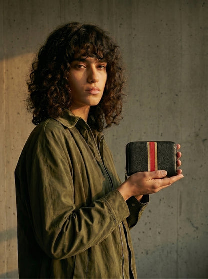 Une jeune femme brune aux cheveux bouclés montre un portefeuille en cuir noir avec des bandes en cuir rouge et doré pâle. 